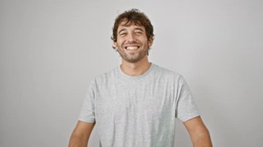 Neşeli genç bir adam spor tişörtü giyiyor, mutlu bir başarı jesti içinde başparmağını kaldırıyor, gülümsüyor. Beyaz duvara yaslanmış, pozitif ışıldıyor..