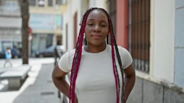Güzel Afrikalı Amerikalı kadın gülüyor, kendine güvenen gülüşü ve günlük tarzıyla şehrin sokaklarına neşe saçıyor, güneşli gökyüzünün altında pozitif enerji yayıyor.