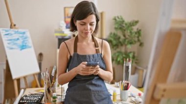 Parlak gözlü genç İspanyol kadın sanatçı, gülümseyen ve kendinden emin, resim yaparken akıllı telefonu ustaca kullanıyor.