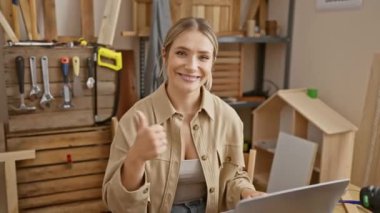 Neşeli genç sarışın kadın, başparmak yukarı, dizüstü bilgisayar, marangozluk atölyesinde çok mutlu.