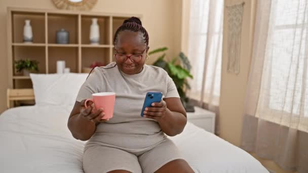 自信的非洲裔美国女人 皮肤舒适 躺在床上休息 喝着早茶 用手机发短信 辫子躺在枕头上 微笑照亮房间 — 图库视频影像