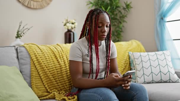 身穿辫子坐在家里沙发上 沉浸在客厅里用智能手机的非洲裔美国女人很不安 — 图库视频影像
