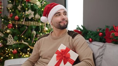 Genç Arap adam elinde hediyeyle evdeki Noel ağacının yanındaki koltukta oturuyor.