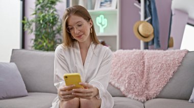 Kanepede oturmuş akıllı telefon kullanan genç sarışın kadın evde gülümsüyor.