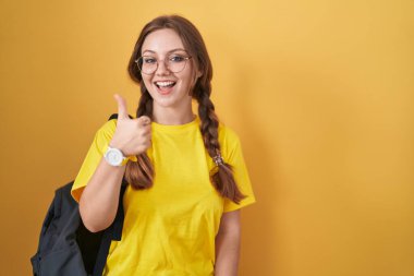 Sarı arka planda öğrenci çantası takan beyaz bir kadın mutlu bir el hareketi yapıyor. Kameraya bakıp başarıyı gösteren ifade. 