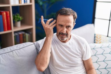 Middle age man listening voice message by smartphone sitting on sofa at home