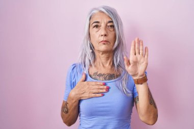 Pembe arka planda duran, elleri göğsünde ve avucunda küfreden, sadakat yemini eden orta yaşlı bir kadın. 