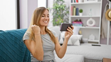 Coşkulu genç sarışın kadın, rahat dairesinde, koltuğunda rahatça otururken tezahürat yapıyor, cep telefonuyla heyecan verici bir video oyunu oynuyor.