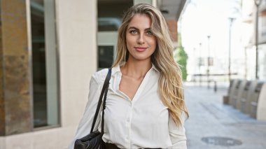 Işıl ışıl, genç, sarışın bir kadın, kendine güveni tam, güneşli bir şehir caddesinde duruyor, şehir ortamının tadını çıkarıyor ve pozitif titreşimler yayıyor..
