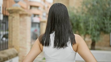 Genç, güzel, İspanyol bir kadın, sıradan, açık havada çekilmiş bir portrede, yeşil bir şehir parkında dikiliyor, arkası da çekici bir saç stilini gözler önüne seriyor..