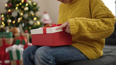 Genç Çinli bir kadın hediye paketini açıyor evde Noel ağacının yanında oturuyor.