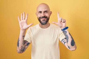 Sarı arka planda dikilen İspanyol dövmeli adam yedinci parmaklarını gösterip işaret ederken kendinden emin ve mutlu gülümsüyor.. 