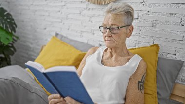 Gri saçlı, son sınıf öğrencisi bir kadın kendini bir kitaba kaptırmış, rahat bir şekilde yatakta dinleniyor, yatak odasının tanıdık sükunetiyle çevrili.