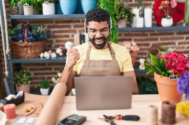 Hispanic man with beard working at florist shop doing video call screaming proud, celebrating victory and success very excited with raised arm 
