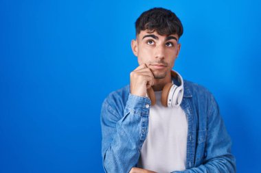 Young hispanic man standing over blue background with hand on chin thinking about question, pensive expression. smiling with thoughtful face. doubt concept. 