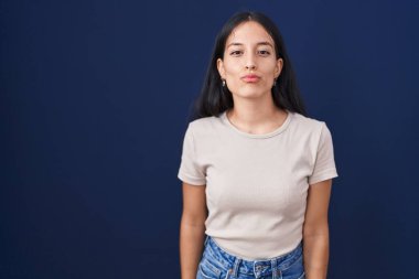 Genç İspanyol kadın mavi arka planda dikilip kameraya bakıyor ve güzel ve seksi bir şekilde yayına öpücük yolluyor. aşk ifadesi. 