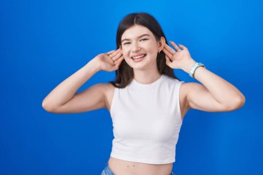 Young caucasian woman standing over blue background smiling pulling ears with fingers, funny gesture. audition problem 