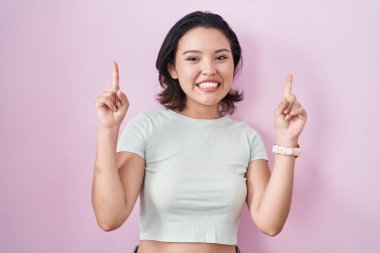 Pembe arka planda duran Latin kökenli genç bir kadın. Şaşırmış, şaşırmış, parmaklarıyla işaret ediyor ve kollarını kaldırıyordu.. 