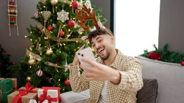 Genç Arap adam akıllı telefonuyla özçekim yapıyor. Evde Noel ağacının yanında oturuyor.