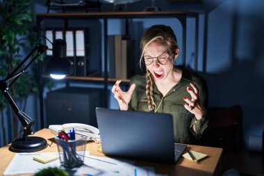 Young blonde woman working at the office at night crazy and mad shouting and yelling with aggressive expression and arms raised. frustration concept. 