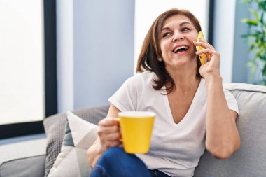 Middle age woman talking on smartphone drinking coffee at home