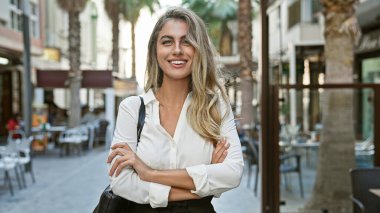 Neşeli bir ifade, neşeli bir gülümseme ve kollarını kavuşturarak duran genç, kendine güvenen bir sarışın kadın. Şehir kahvecisinin terasında rahat bir poz.