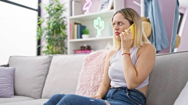 Genç sarışın kadın akıllı telefonda konuşuyor. Evdeki kanepede oturuyor.