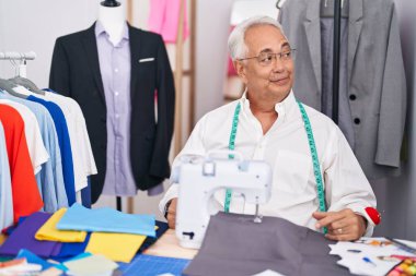 Orta yaşlı bir adam, gri saç terzisi dikiş makinesi kullanıyor. Gülümsüyor ve başka tarafa bakıyor.. 