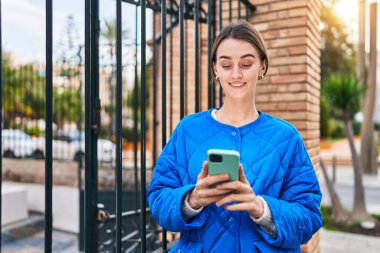 Beyaz bir kadın sokakta akıllı telefon kullanarak gülümsüyor.