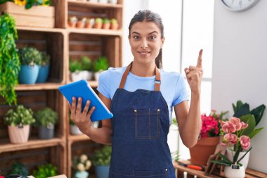 Çiçekçi dükkanında çalışan esmer bir kadın elinde tabletle gülümseyen bir fikirle ya da soru sorarak mutlu bir yüzle işaret ediyor. 
