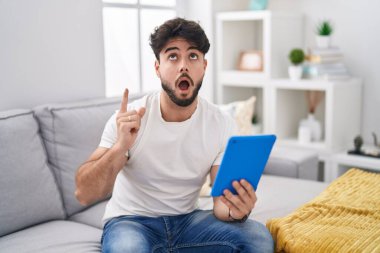 Kanepeye oturmuş dokunmatik ped kullanan İspanyol sakallı adam şaşırmış ve şaşırmış bir şekilde yukarı baktı ve parmaklarıyla işaret etti ve kolunu kaldırdı.. 