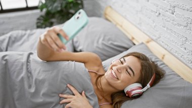 Neşeli genç İspanyol kadın rahat yatağında uzanıyor, mutlu bir dijital sohbete dalmış. Kulaklık takıyor, güzel yatak odasında akıllı telefonunu kullanıyor..