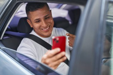 Genç İspanyol adam akıllı telefonuyla video görüşmesi yapıyor. Arabada gülümsüyor. Elinle ve parmağınla mutlu mesut işaret ediyor. 