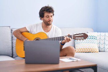 Latin kökenli genç bir adam evdeki kanepede oturup online gitar dersi alıyor.
