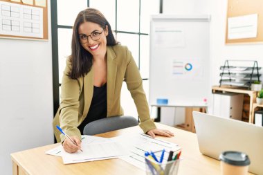 Young beautiful hispanic woman business worker writing on document standing at office