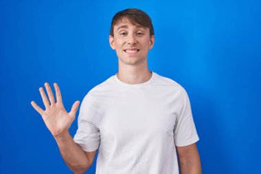 Mavi arka planda duran beyaz tenli sarışın adam beş numarayı gösterip işaret ederken kendinden emin ve mutlu bir şekilde gülümsüyor.. 