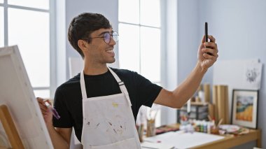 Gülümseyen genç İspanyol ressam akıllı telefonuyla fevkalade bir özçekim fotoğrafı çekiyor. Bir elinde tuval, diğer elinde boya fırçası.!
