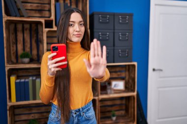 Genç esmer kadın akıllı telefonu açık elle kullanıyor dur işareti yapıyor ciddi ve kendinden emin bir ifadeyle, savunma hareketi yapıyor. 