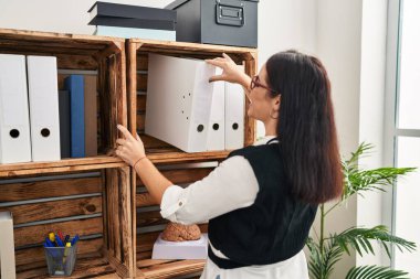 Young beautiful hispanic woman business worker holding documents of shelving at office