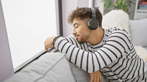 年轻的阿拉伯男子喜欢自己的声音避难所 在沙发上放松 全神贯注于耳机的音响技术 在家里听音乐 — 图库照片