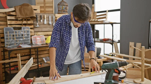 Handsome young arab man, a professional carpenter, concentrated on drawing a mark on a wood plank, fully engrossed in his carpentry work inside the workshop