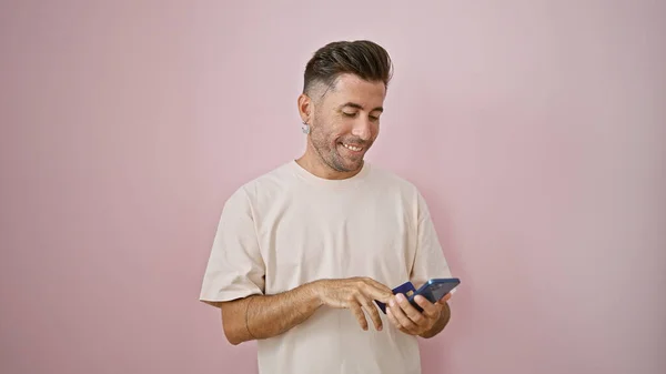 Smiling young hispanic man confidently tapping into online shopping over isolated pink background, smartphone in hand, his credit card making business and finance a casual affair