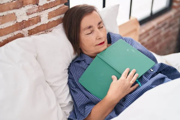 Middle age woman holding book lying on sofa sleeping at bedroom