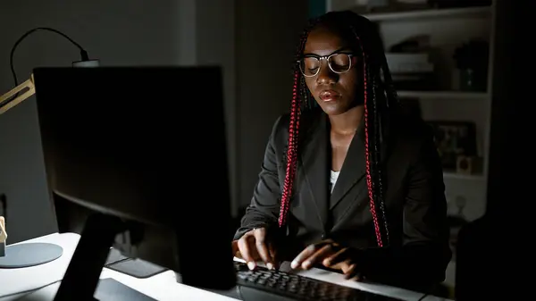 Focused african american woman boss governs her business empire night and day, navigating success from her workplace desk, mastering the internet on her office computer screen