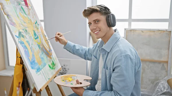 Confident handsome young caucasian man joyfully listening to music while painting, unleashing his creativity at indoor art studio