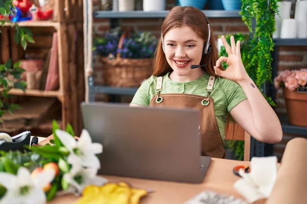 Redhead woman working at florist shop doing video call doing ok sign with fingers, smiling friendly gesturing excellent symbol