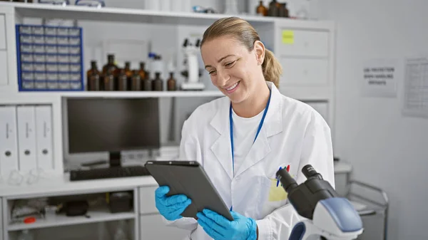 ブロンドの女性科学者は 革新的な医療技術に従事しながら 彼女のタッチパッドを楽しんで 若くて自信を持って研究室で優れた笑顔 — ストック写真