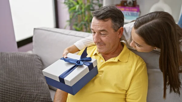 At home, a surprised father and daughter smiling together, sitting on the sofa with a gift in a loving hispanic family relaxing living room.