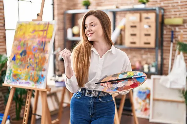 Young caucasian woman at art studio holding palette pointing thumb up to the side smiling happy with open mouth