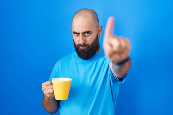 Young hispanic man with beard and tattoos drinking a cup of coffee pointing with finger up and angry expression, showing no gesture
